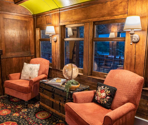 Cozy interior of a train car with two armchairs, a globe, and wooden paneling, illuminated by wall sconces.